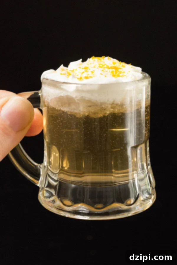 A hand holds up a shot glass filled with a butterbeer shot, topped with whipped cream and gold sprinkles.