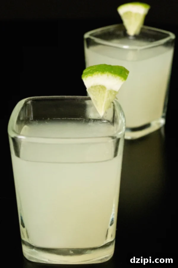 Zesty Lime Shooters 4 One perfectly focused lime drop shot in a square glass, with a blurred background showing another shot glass and limes, all on a black surface.