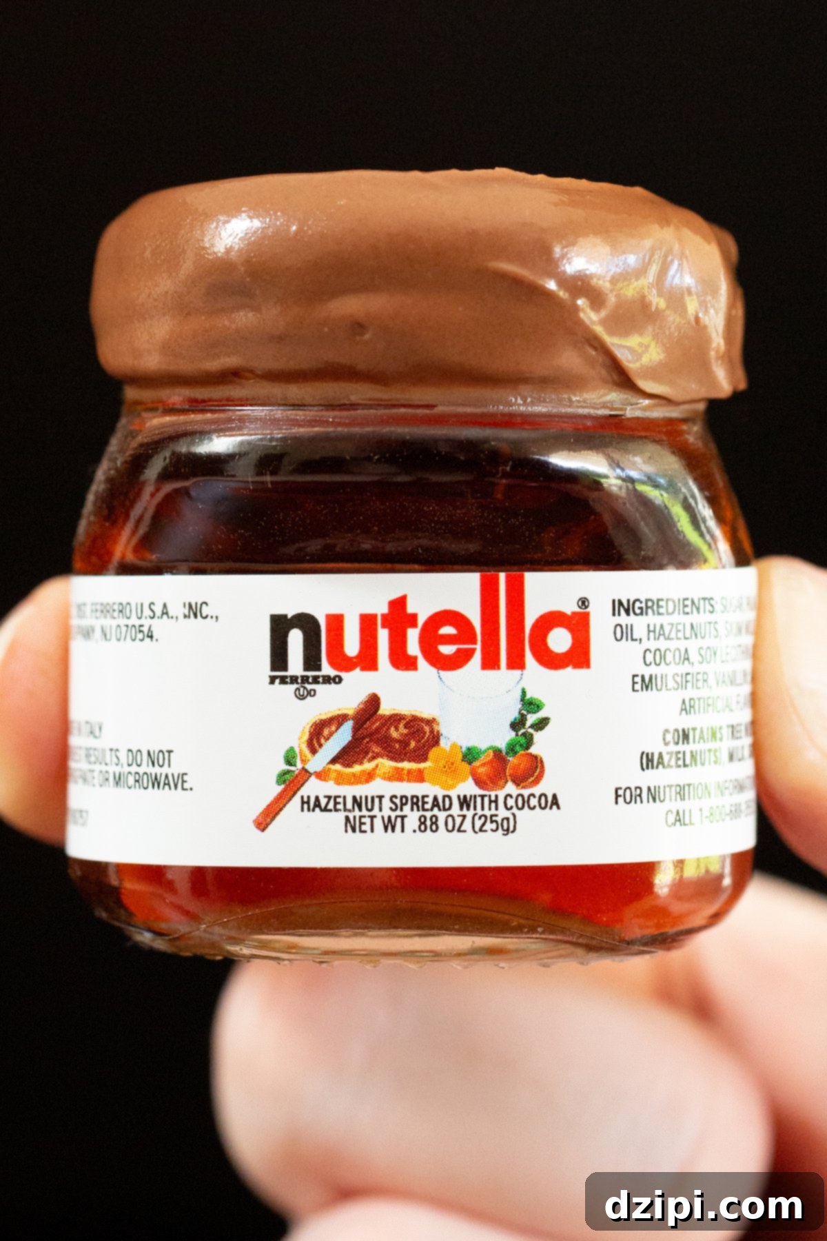 A thumb and index finger hold up a mini Nutella jar that's filled with a Nutella shot. The rim has been dipped in the hazelnut spread.