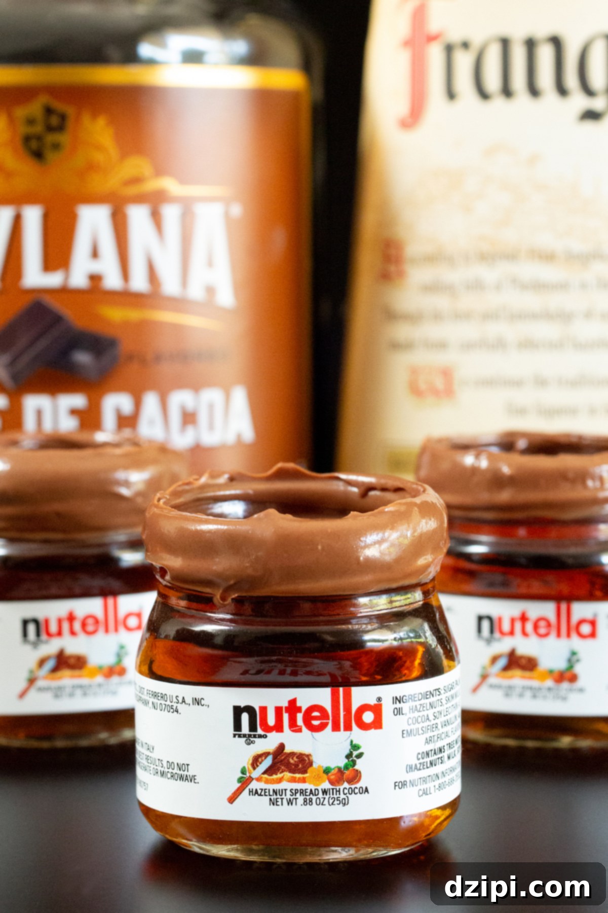 Three miniature Nutella jars with Nutella shots in them in front of creme de cacao and Fragelico liqueur bottles.