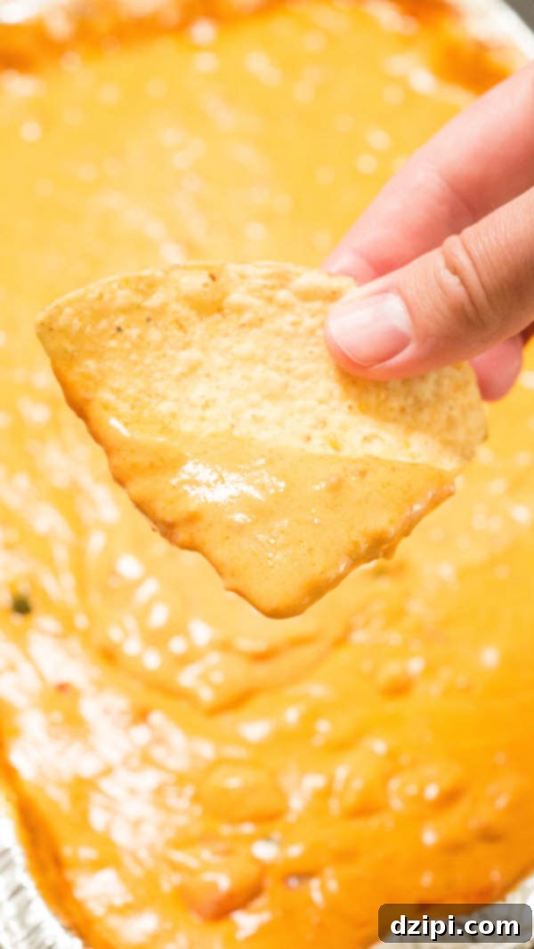 Close-up of a finger holding a tortilla chip that has been dipped in rich, creamy smoked queso, ready to be enjoyed.