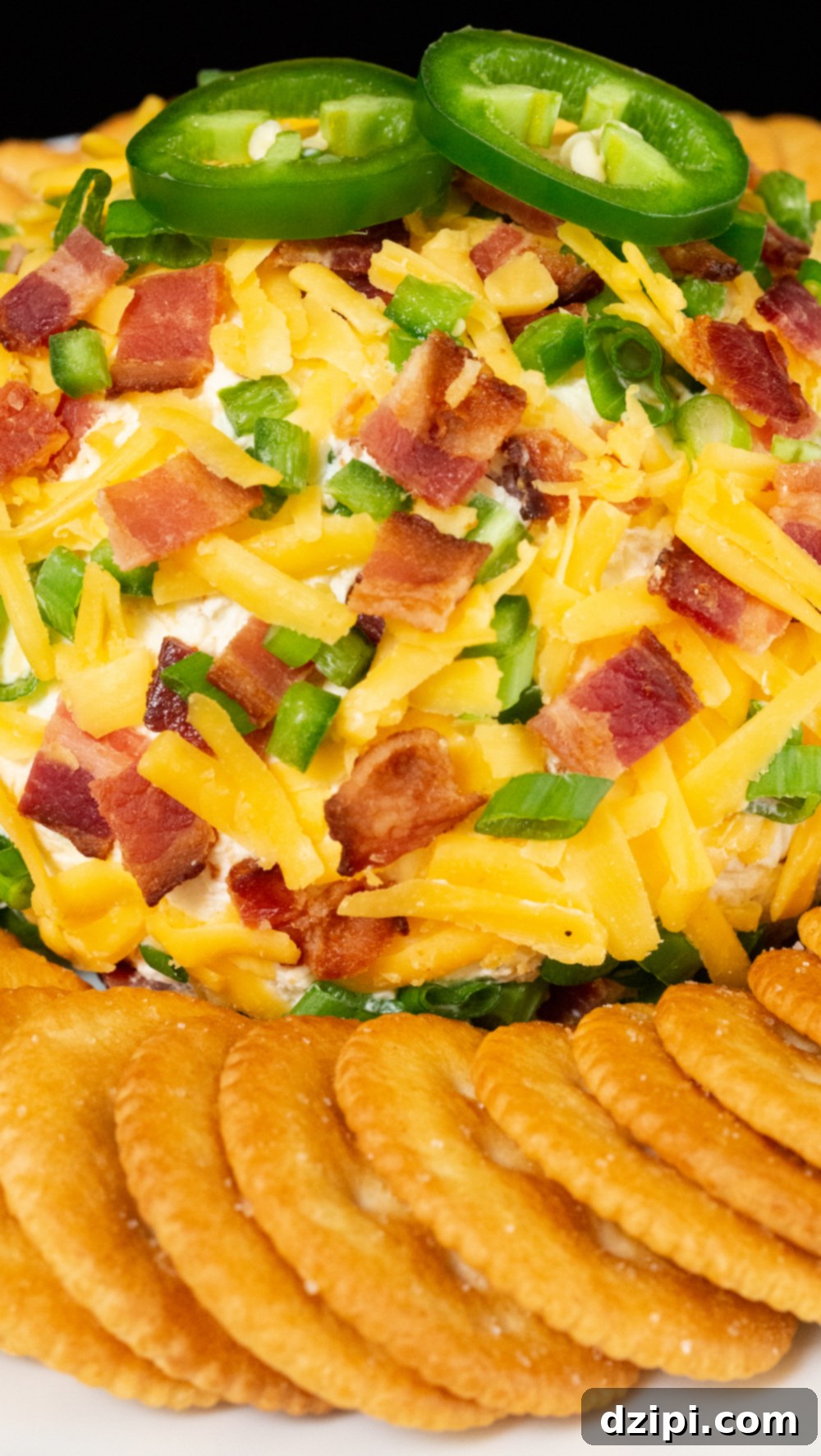 A cheese ball covered in shredded cheddar cheese, chopped, bacon, and chives, sits on the middle of a white plate surrounded by ritz crackers