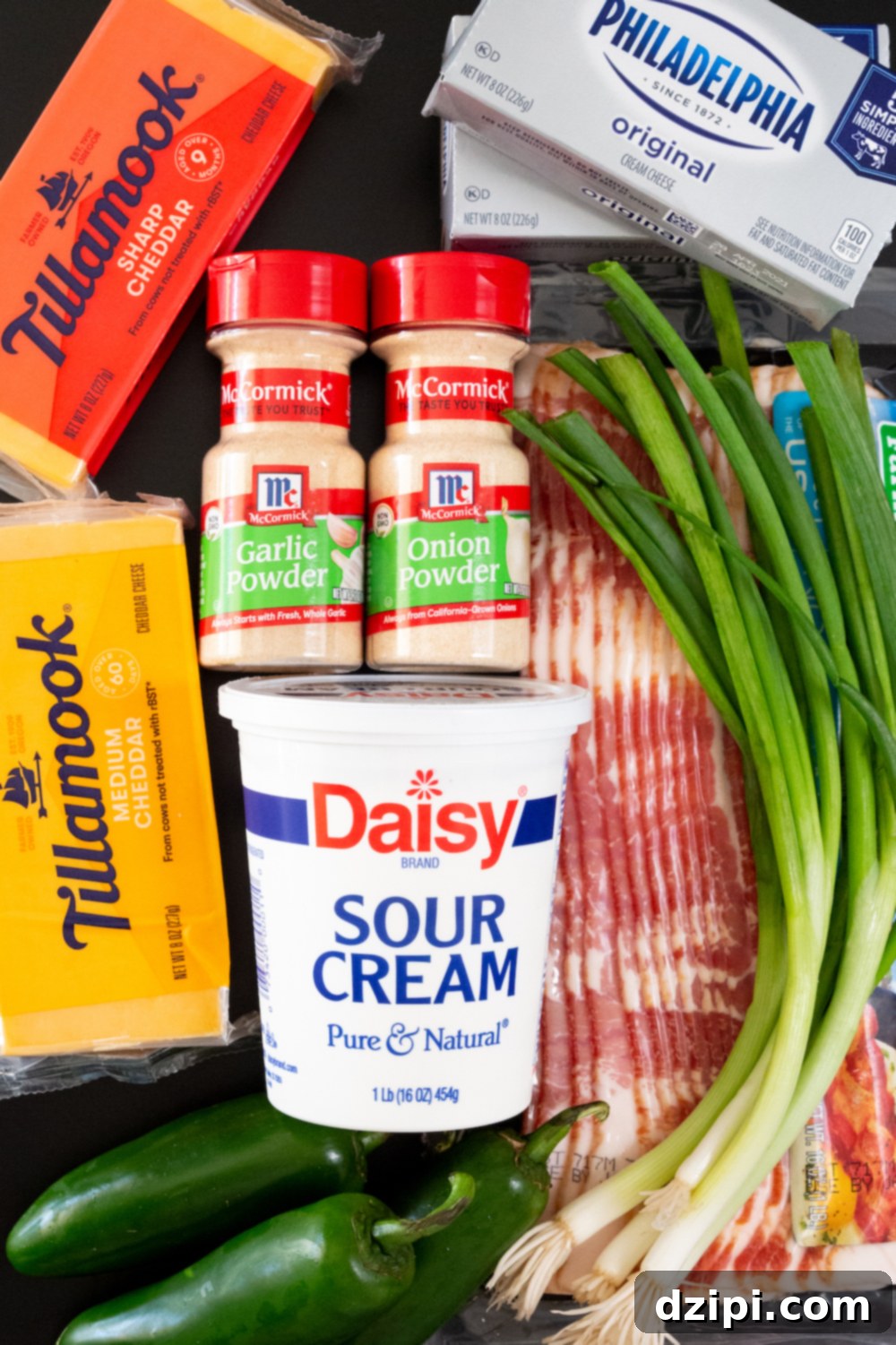 All the ingredients for a jalapeno popper cheese ball lay on a black table (2 blocks of cheddar cheese, cream cheese, onion powder, garlic powder, bacon, sour cream, green onions, and three fresh jalapenos).