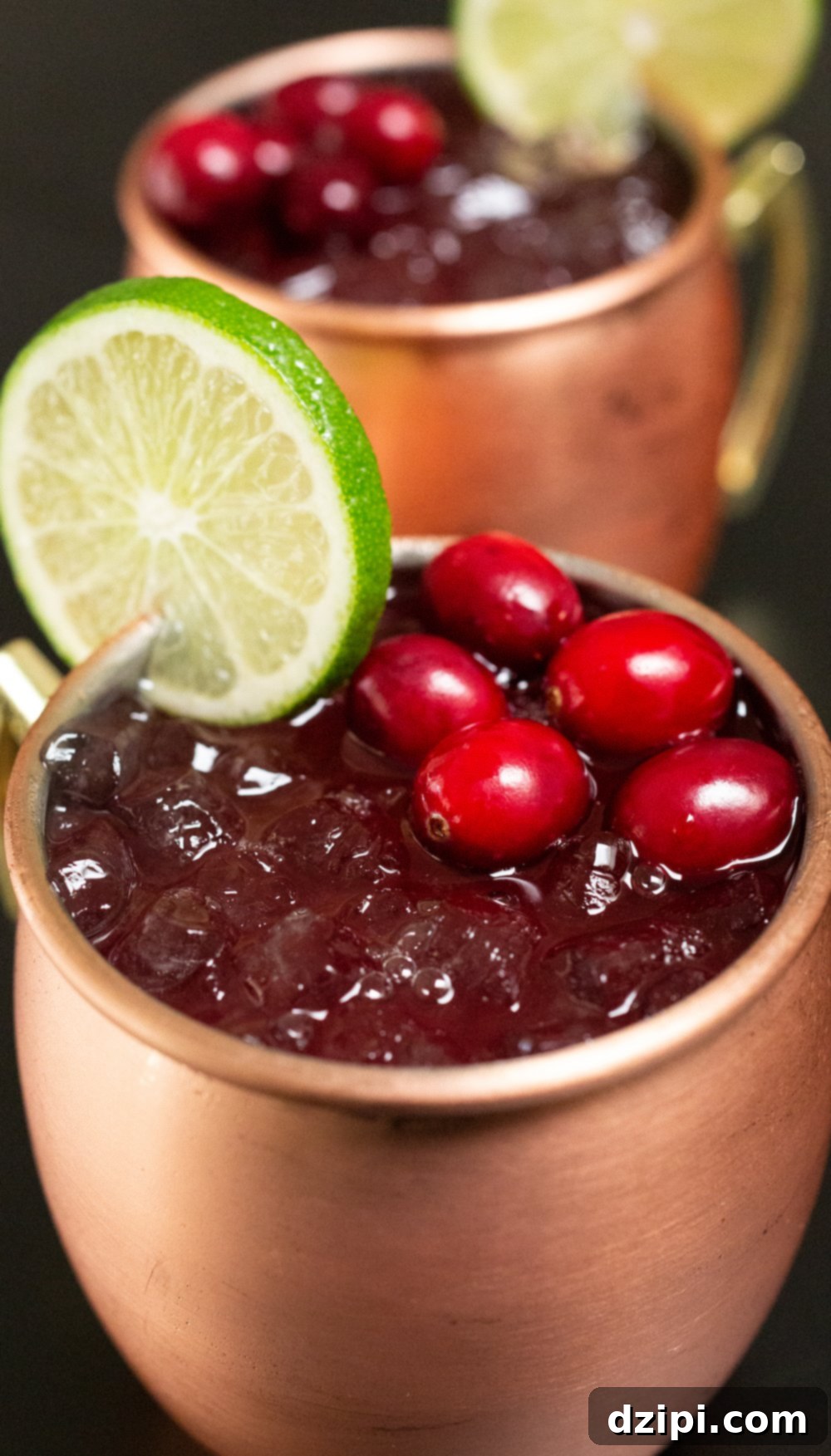 Two glistening copper mugs brimming with festive cranberry Moscow Mules, garnished with a vibrant lime wheel and fresh cranberries, are artfully arranged against a sleek black background.
