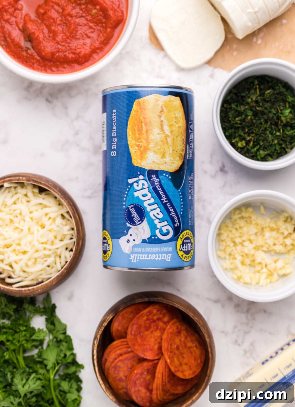 An overhead shot showcasing all the fresh ingredients ready for the biscuit pizza casserole: large canned Grands! Biscuits, a bowl of shredded mozzarella, fresh mozzarella slices, a stack of pepperoni, a jar of rich pizza sauce, fresh parsley, and a block of butter with minced garlic.