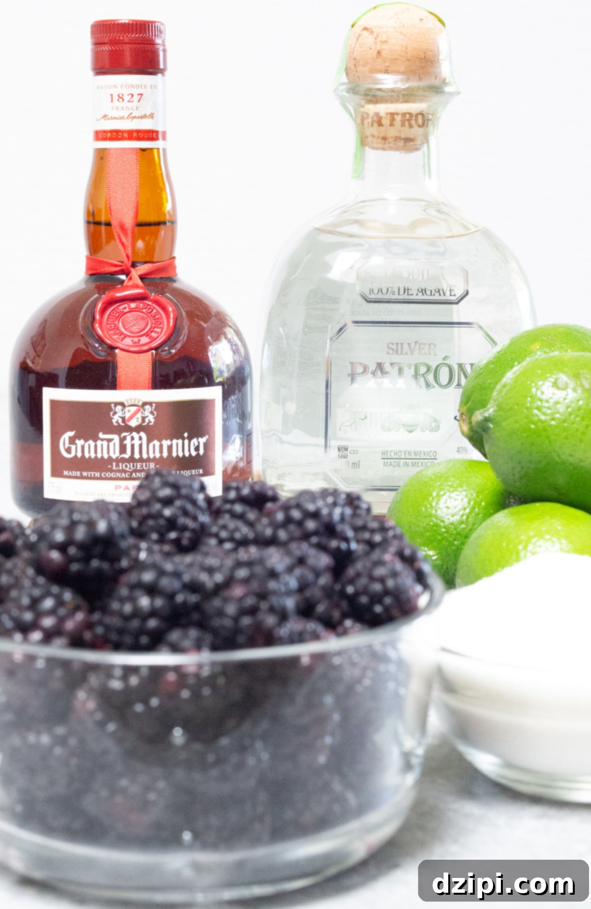 A flat lay of fresh ingredients for blackberry margaritas: bottles of Grand Marnier and Patron tequila, a pile of ripe blackberries, several bright green limes, and a small bowl of sugar. All arranged on a white background, highlighting the vibrant colors and natural freshness of the components.