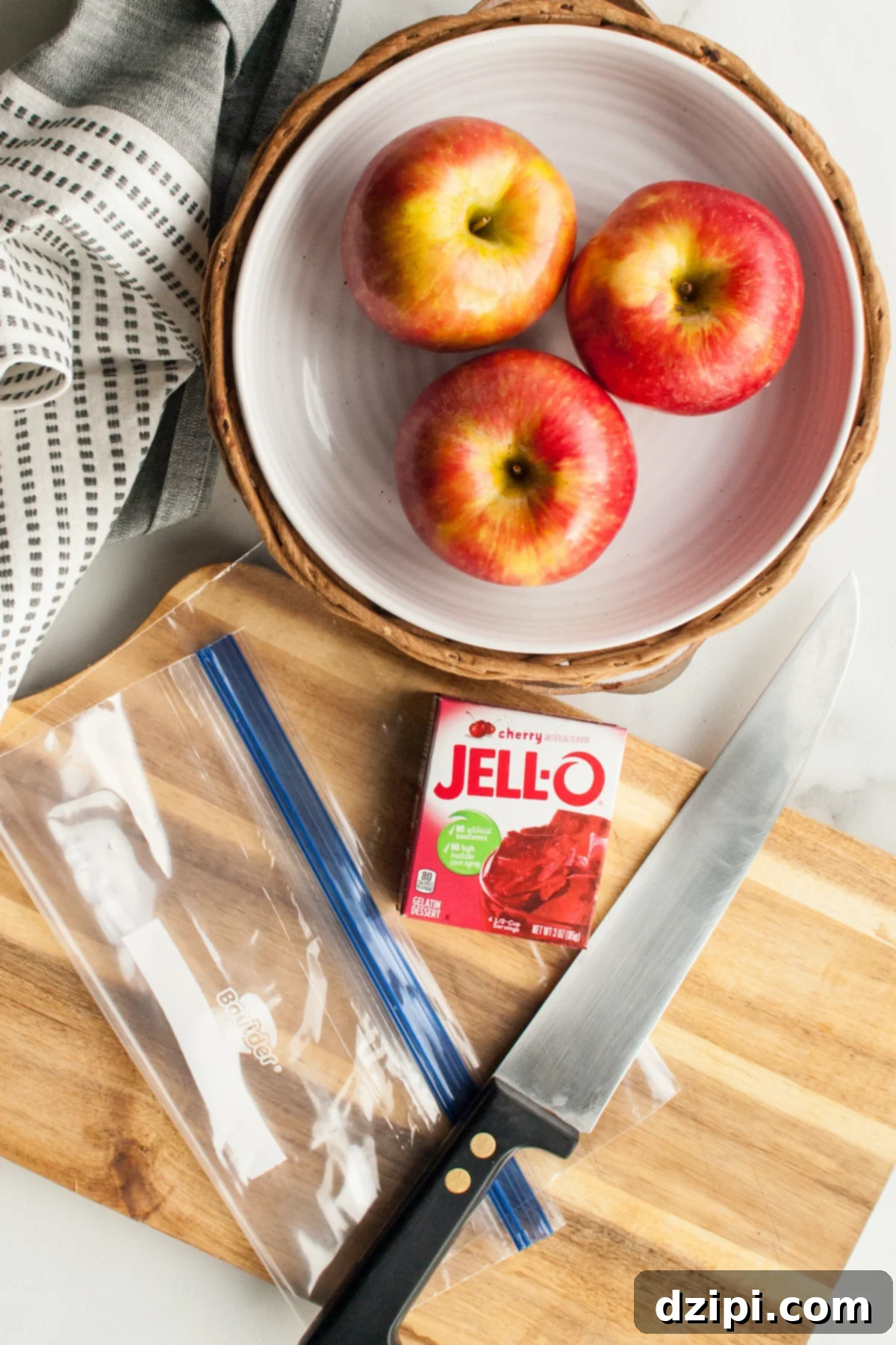 Overhead view of jello apple ingredients (3 apples, 1 box of jello powder) sitting next to a knife, ziploc bag, and a cutting board.