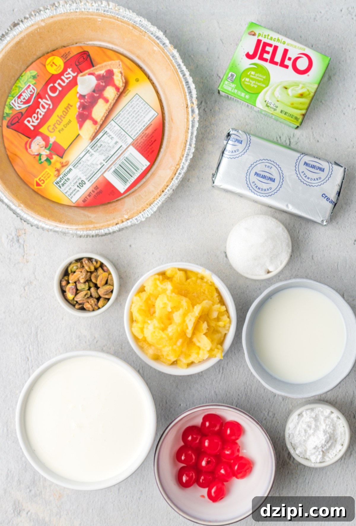 Emerald Dream Pie 4 An overhead shot showcasing all the key ingredients laid out for pistachio pie: a ready-made graham cracker crust, a box of Jello Pistachio pudding mix, a block of cream cheese, a can of crushed pineapple, granulated sugar, maraschino cherries, chopped pistachios, milk, and heavy cream.