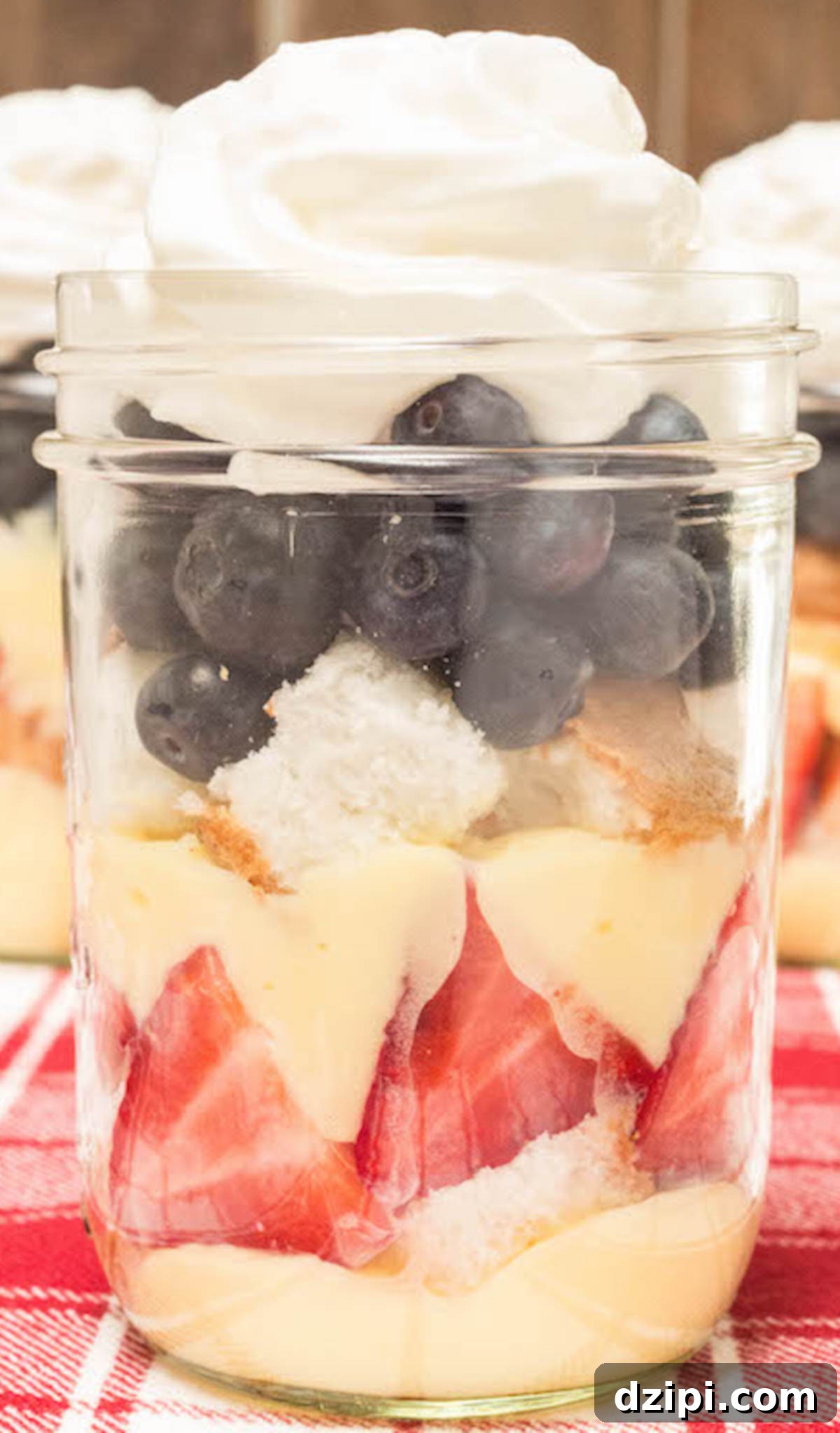 A small mason jar elegantly layered with pudding, sliced strawberries, angel food cake cubes, blueberries, and whipped cream, resting on a festive red picnic tablecloth.