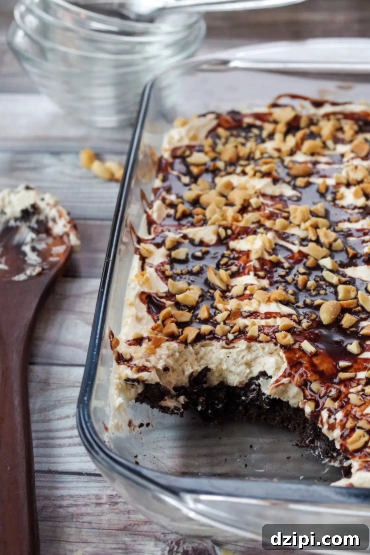 A glass casserole dish filled with a Drumstick Dessert, showcasing a chocolate cake base topped with whipped cream, drizzled with chocolate sauce, and garnished with crushed peanuts.