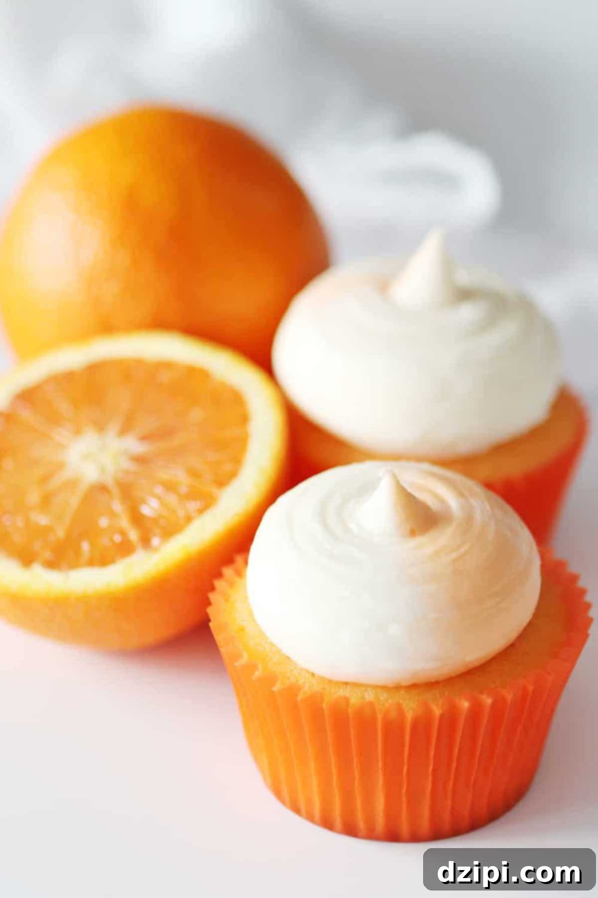 Two fluffy orange creamsicle cupcakes, topped with pristine white frosting, sit beside a halved orange on a light background, highlighting their citrusy appeal.