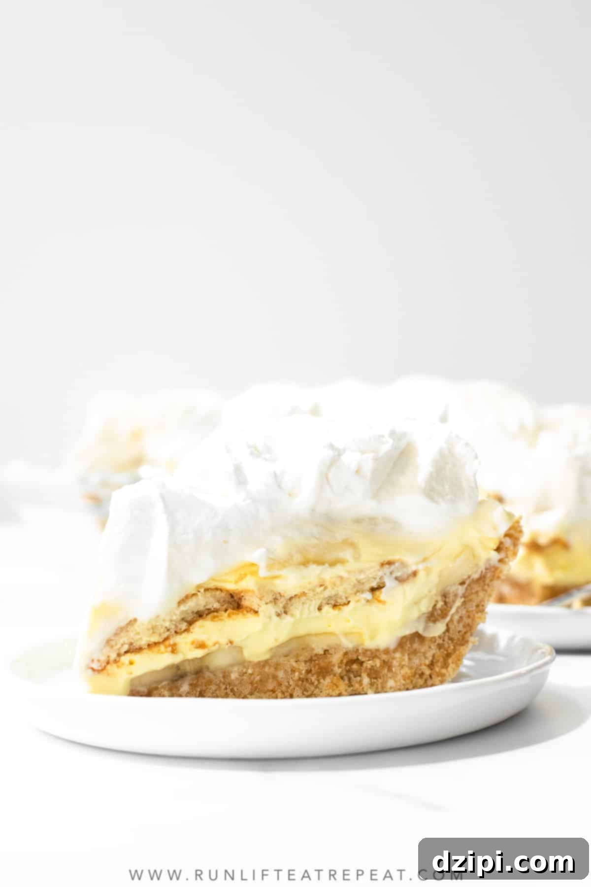 A single slice of creamy Banana Pudding Pie, with its distinctive layers of cookies, bananas, and whipped cream, sits on a white serving plate in front of the full pie.