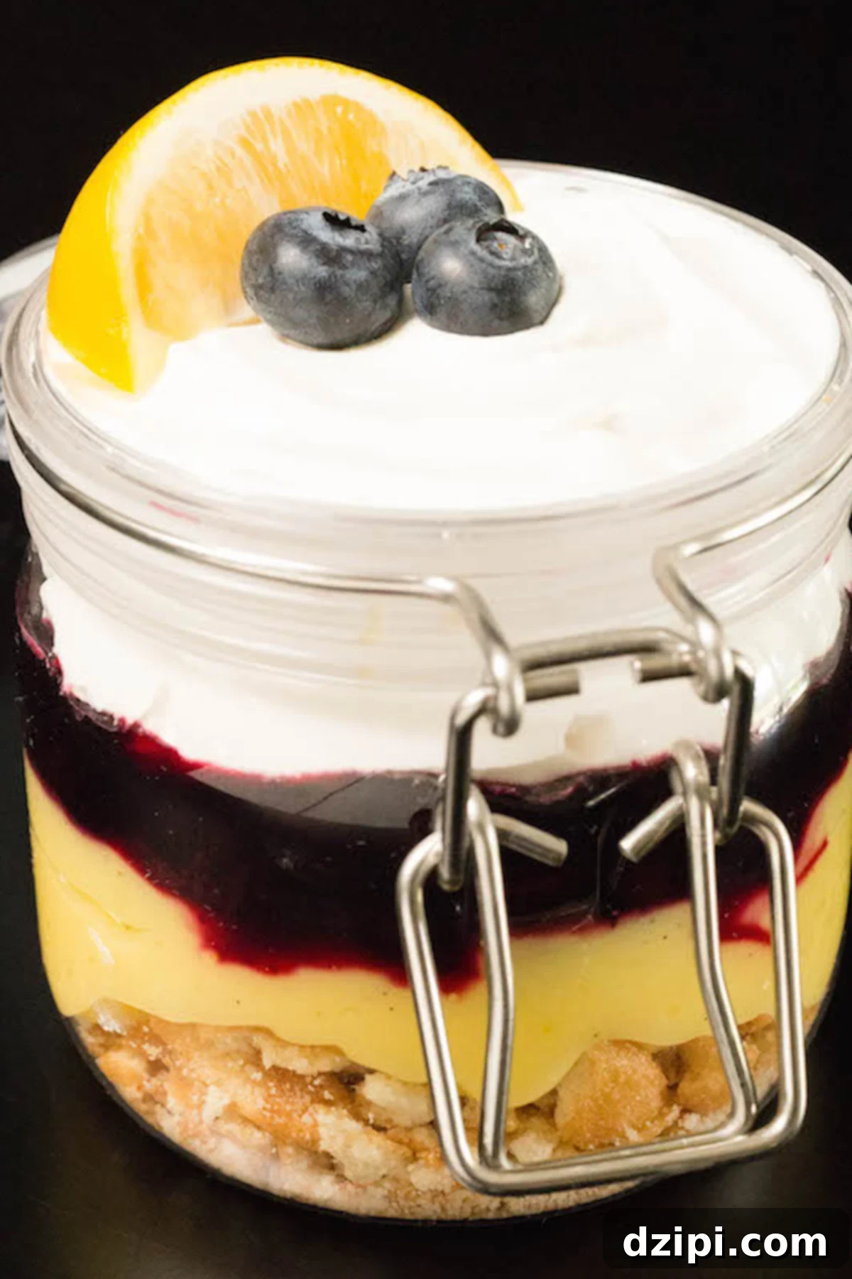 A small, elegant glass jar filled with beautiful layers of Nilla Wafers, bright lemon curd, sweet blueberry compote, and airy whipped cream, set against a dark background.