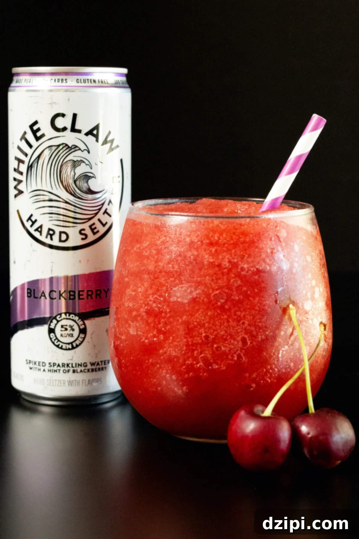 A stemless wine glass filled with a red white claw slushie sits in front of a White Claw can on a black background.