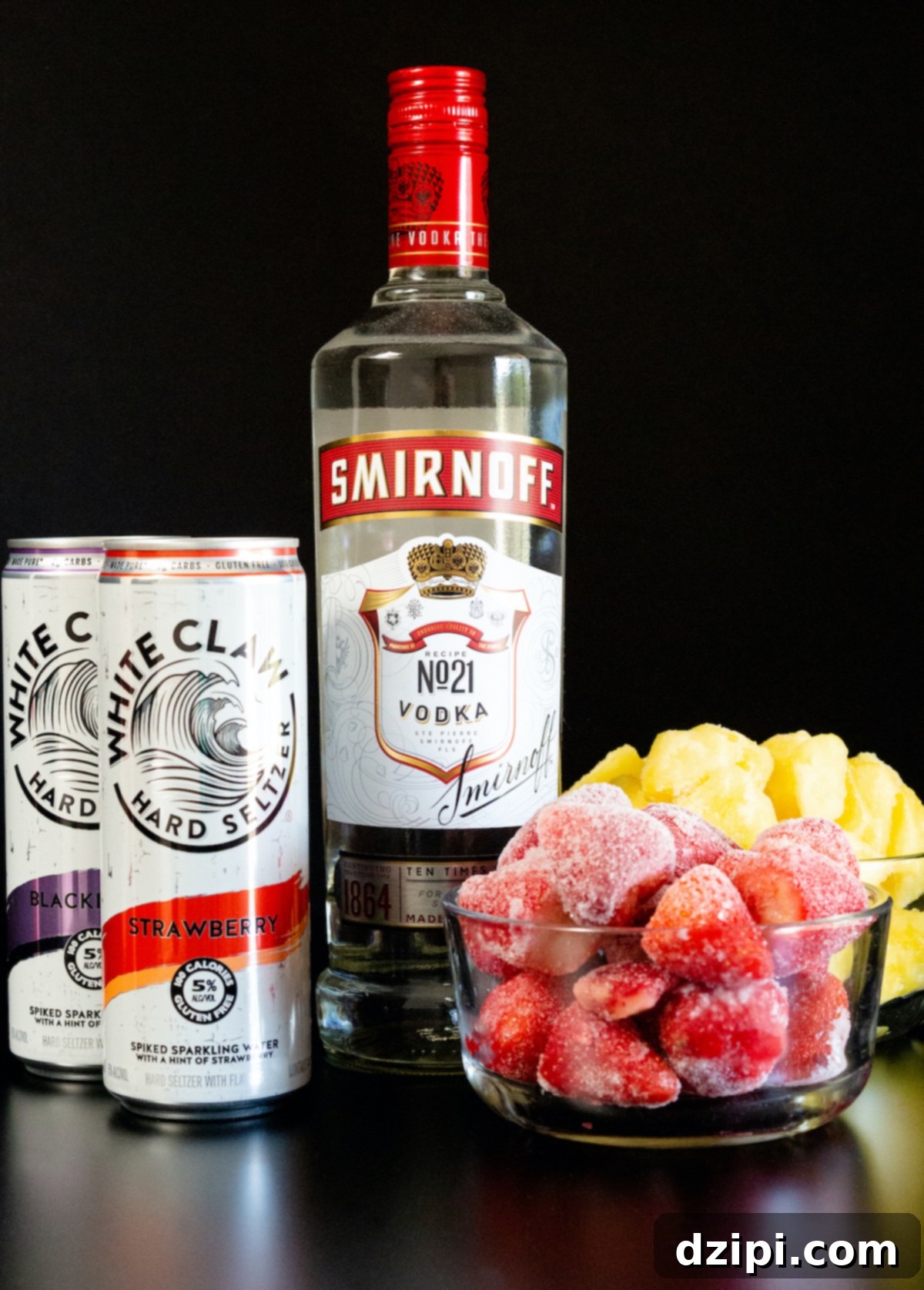 All the ingredients to make a white claw slushy sit on a black background. A bottle of smirnoff vodka, white claw cans, and frozen fruit.