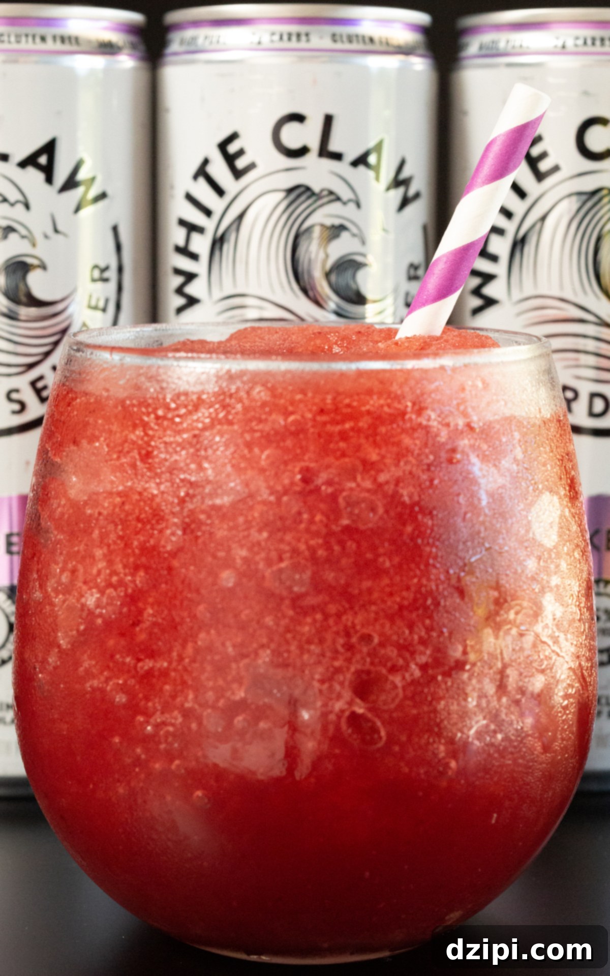 A close up of a stemless wine class filled with a red slushie in front of three cans of White Claw hard seltzer.