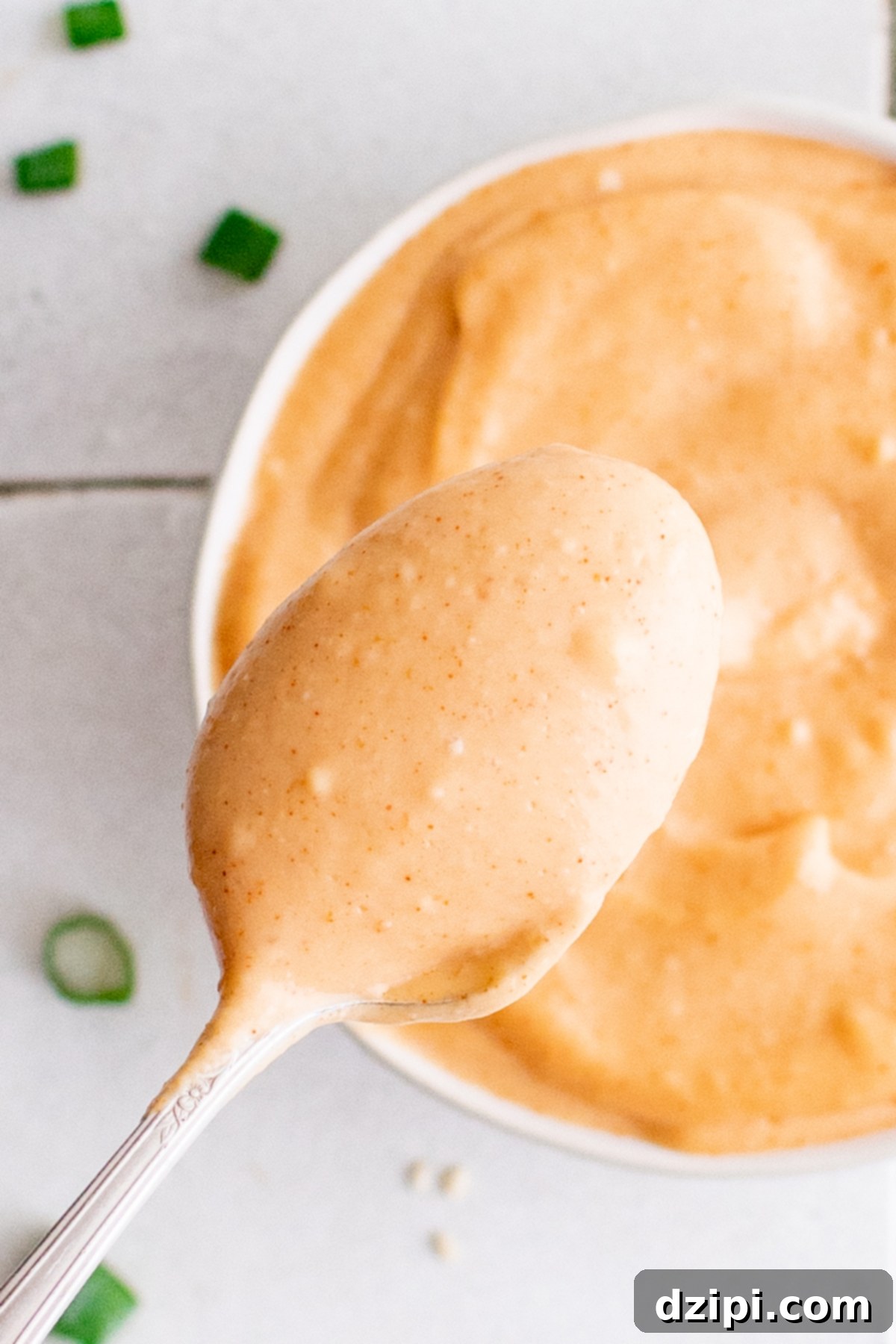 An inviting overhead perspective of a white ceramic bowl generously filled with creamy Yum Yum sauce. A spoon is poised, holding a dollop of the rich sauce just above the bowl, ready to drizzle over your favorite dish.