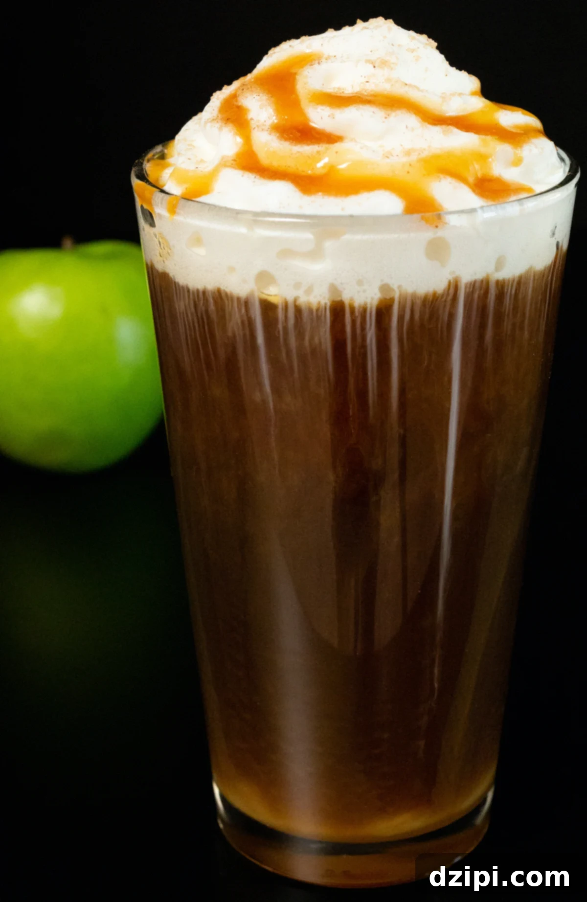 A tall glass of Caramel Apple Cold Brew topped with whipped cream and caramel sauce, next to a vibrant green apple on a dark background, evoking the essence of autumn.