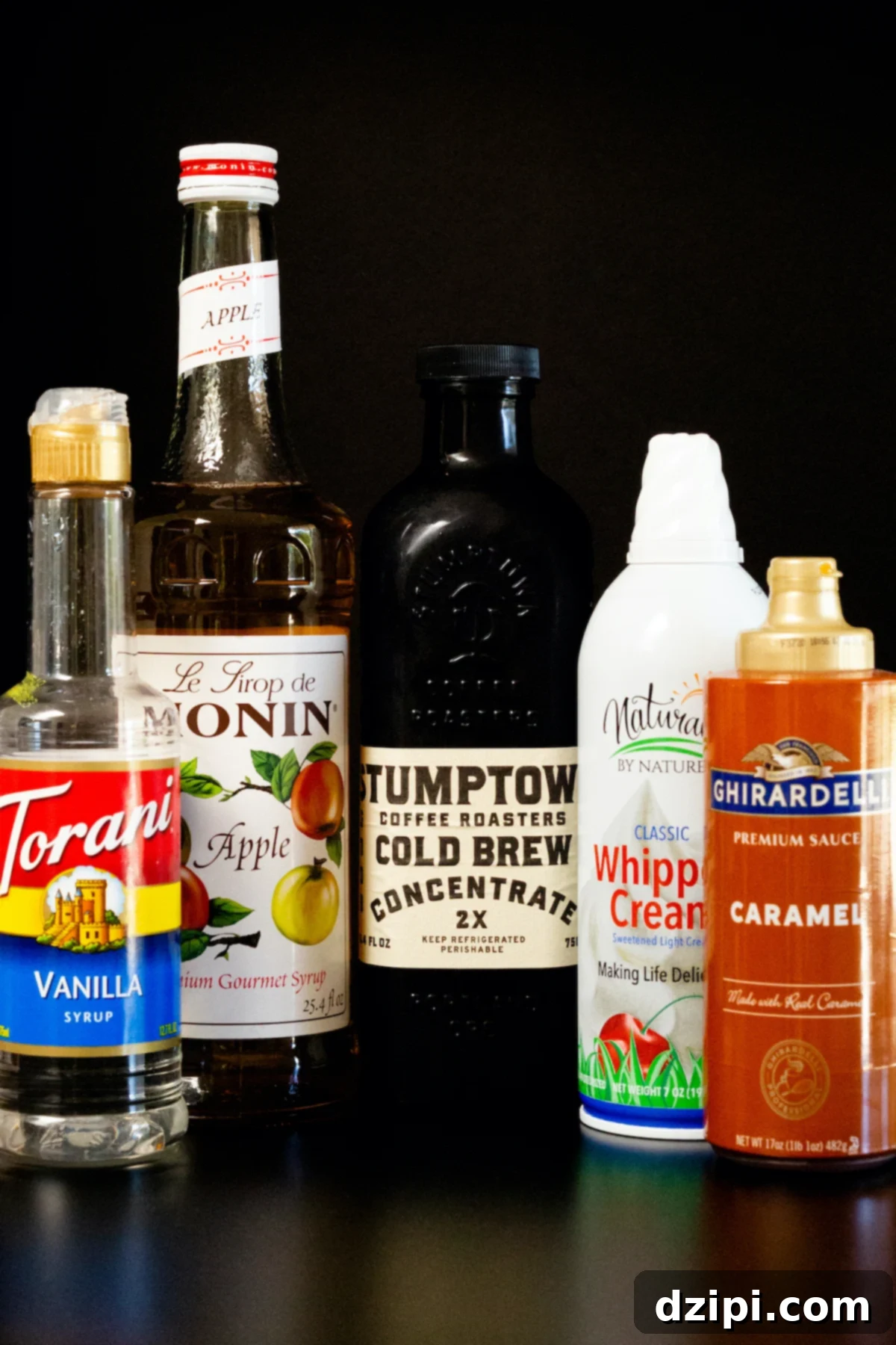 All the necessary ingredients for a delicious Caramel Apple Cold Brew are laid out on a dark surface: bottled cold brew coffee, apple syrup, vanilla syrup, caramel sauce, and whipped cream, ready for assembly.