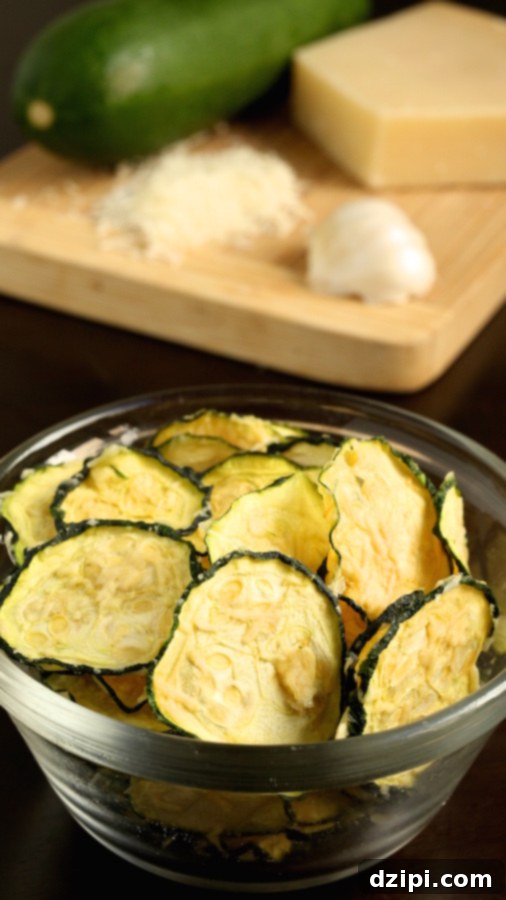 A small glass bowl of Zucchini Chips sits in front of a cutting board with the ingredients (raw zucchini, shredded parmesan, and garlic).