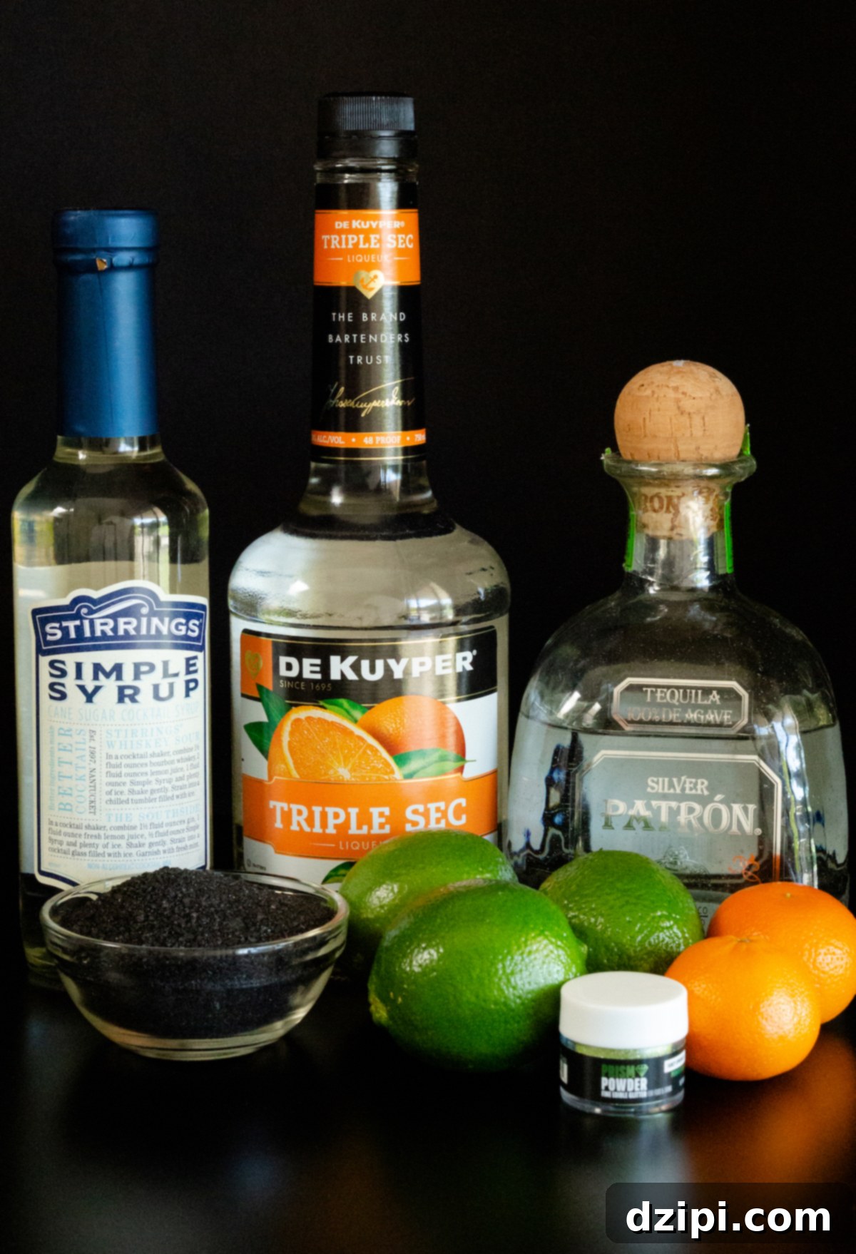 All the ingredients to make halloween margaritas sit on a black background. Simple syrup, triple sec, tequila, lava salt, limes, and small mandarin oranges.