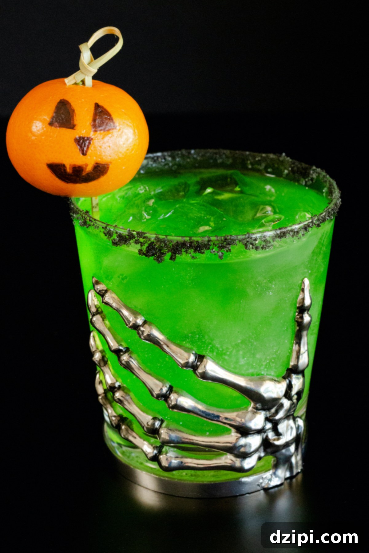 A lowball glass with a skeleton hand on the front is filled with a bright green margarita. There is a black salt rim and jack o lantern garnish.