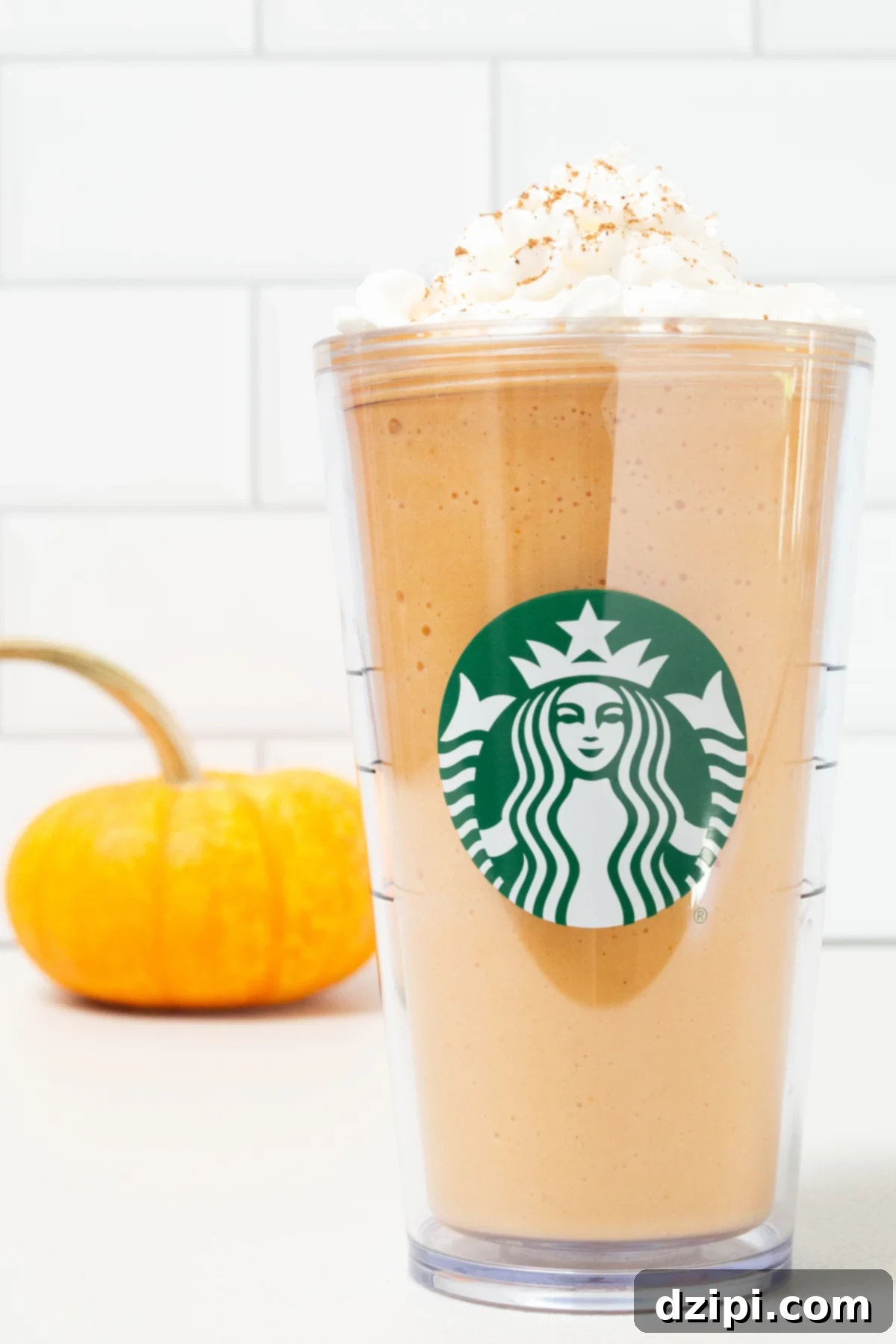 A tall plastic Starbucks tumbler is filled with pumpkin spice frappuccino that's been topped with whipped cream and pumpkin pie spice. A small orange pumpkin is out of focus in the background.