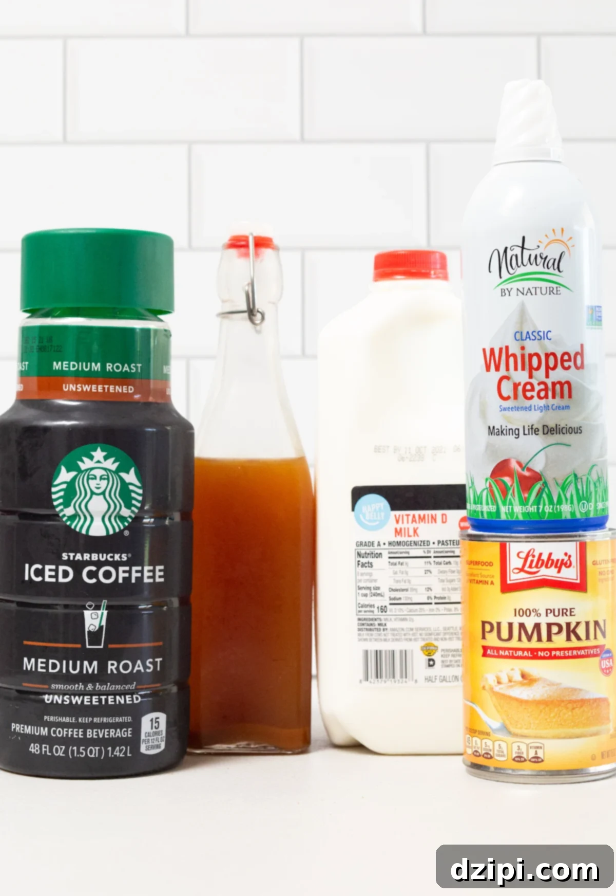 The ingredients to make a pumpkin spice frappuccino sit on a white background - iced coffee, pumpkin spice syrup, whole milk, canned pumpkin, and whipped cream