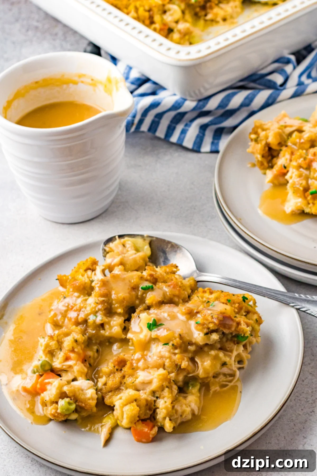 A white plate holds a generous serving of Turkey Stuffing Casserole, with a gravy boat and the remaining casserole in a baking dish visible in the soft-focused background, ready for serving.