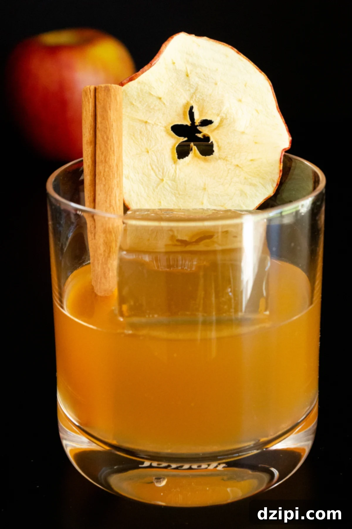 A lowball glass has been filled with a brown Apple Old Fashioned and a large clear block of ice. It is garnished with a dehydrated apple slice and a cinnamon stick, evoking a warm, autumnal feel.
