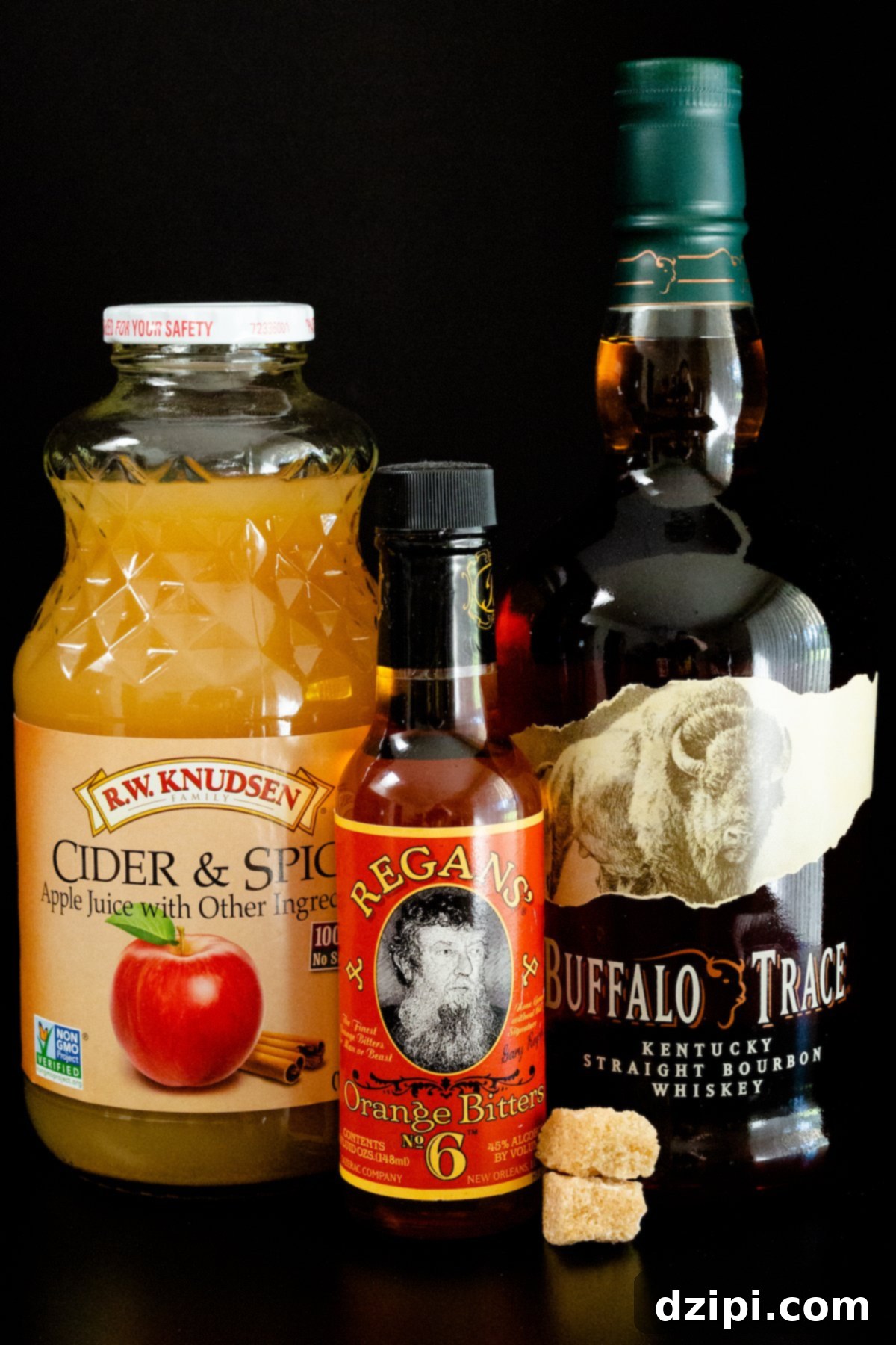All the ingredients to make an apple old fashioned sit on a black background - a bottle of apple cider, bitter, bourbon, and two cubes of brown sugar. The scene is tastefully arranged, highlighting each component.