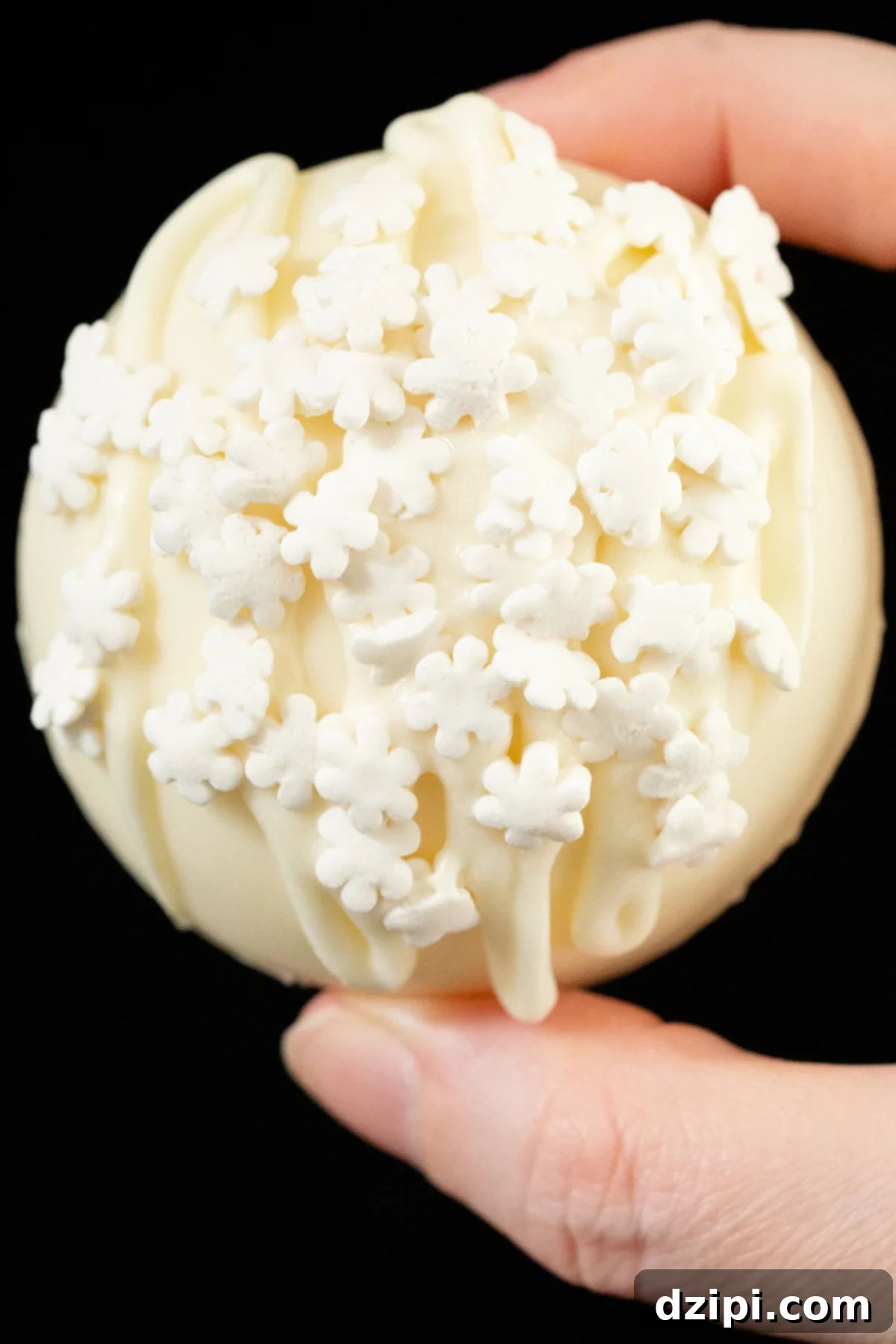 Fingers hold a white hot chocolate bomb that has been drizzled with white chocolate and garnished with white snowflake sprinkles.