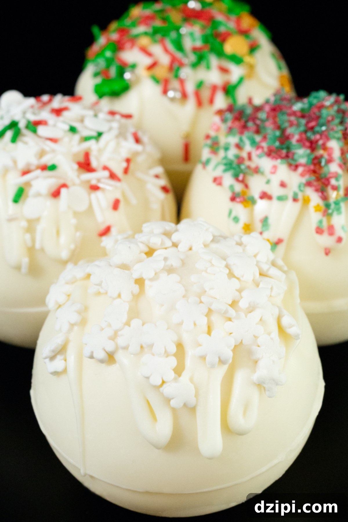 4 white hot chocolate bombs sit on a black background. They are adorned with drizzled white chocolate at various Christmas colored sprinkles.