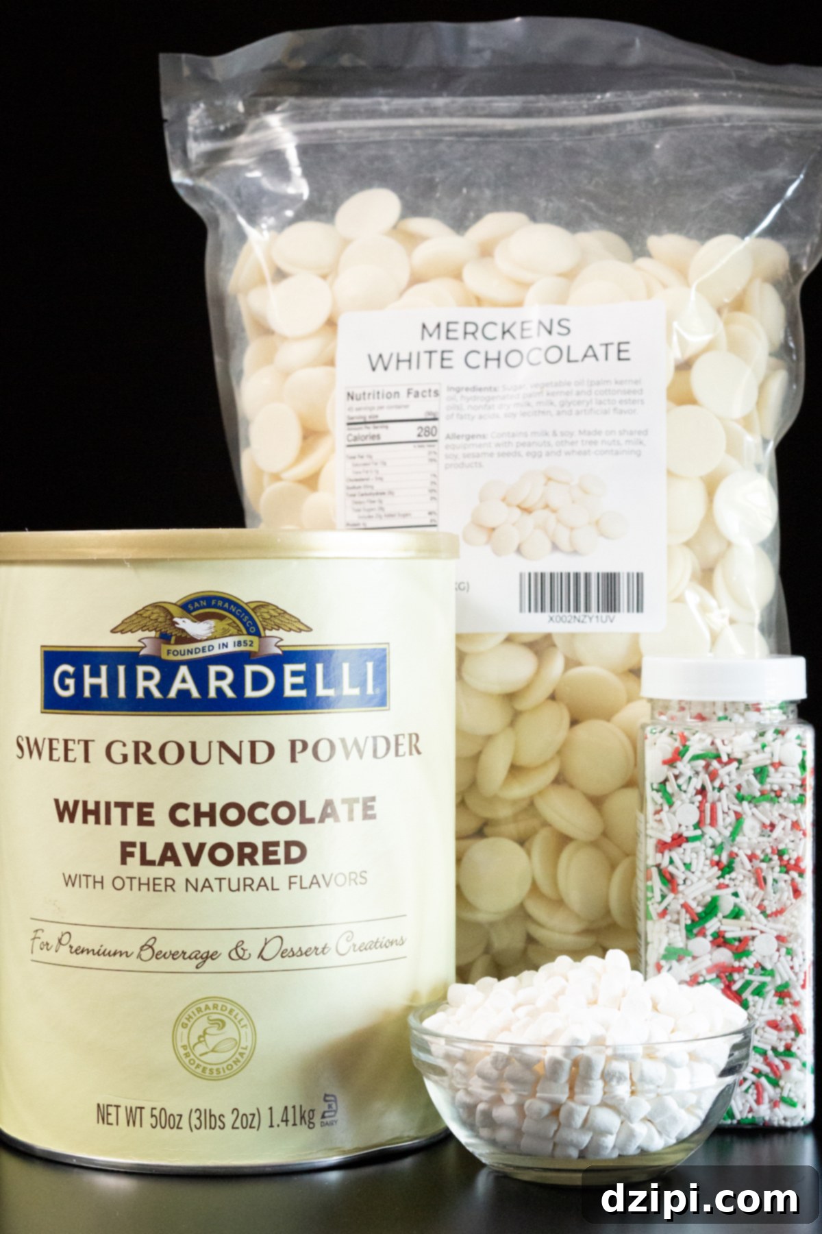 All the ingredients to make white hot chocolate bombs sit on a black background - a tub of Ghirardelli white chocolate powder, dehydrated marshmallows, white chocolate melts, and sprinkles.