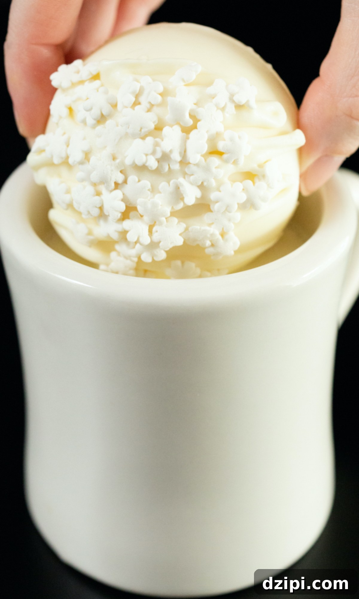 A hand is dropping a white hot chocolate bomb into a white ceramic coffee mug that has been filled with hot milk.