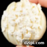 Fingers hold a white hot chocolate bomb that has been drizzled with white chocolate and garnished with white snowflake sprinkles.