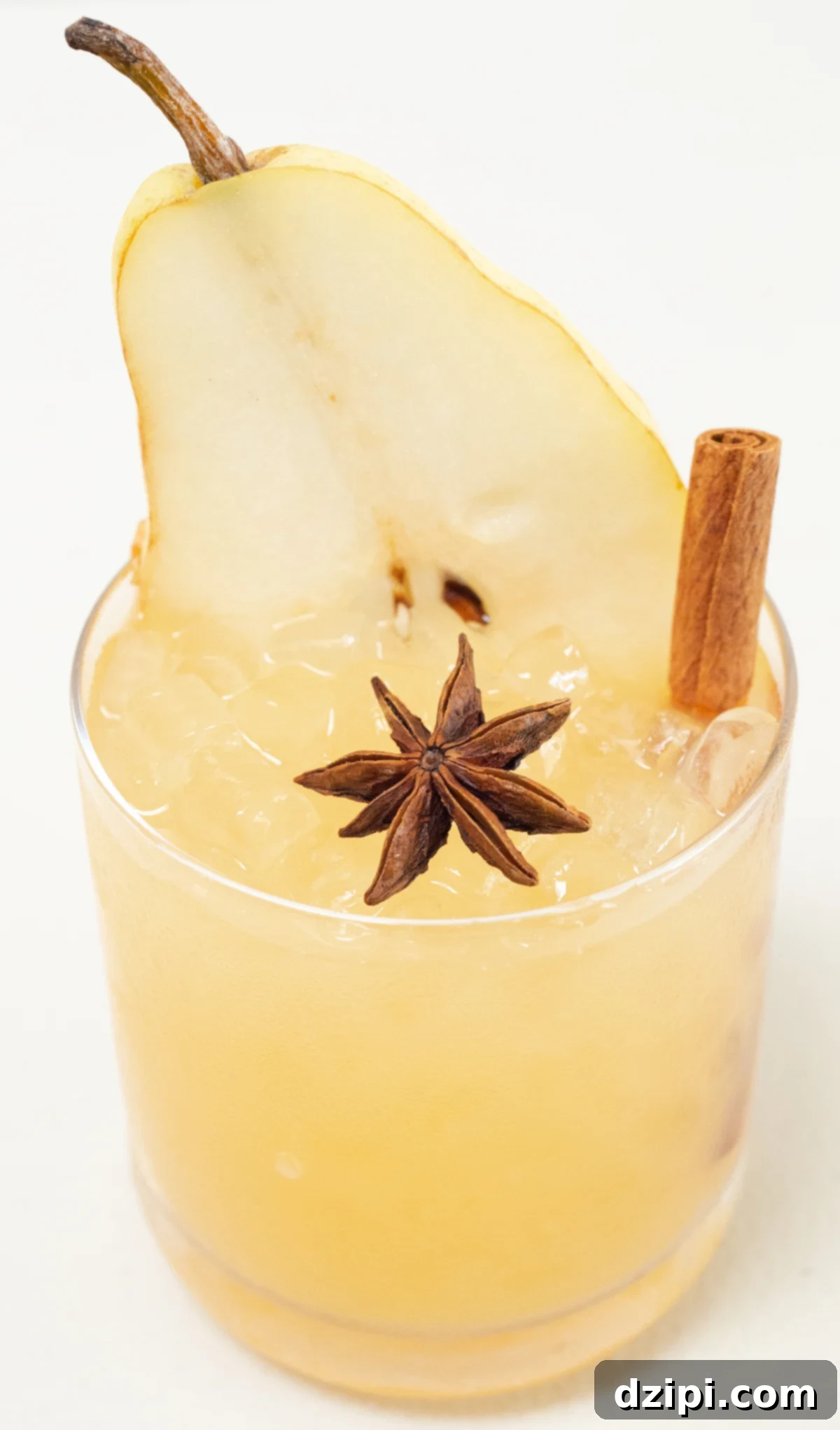 A single lowball glass filled with pebble ice and a vibrant yellow Thanksgiving cocktail, elegantly garnished with a fresh pear slice, aromatic star anise, and a rustic cinnamon stick. The setting evokes warmth and autumn cheer.