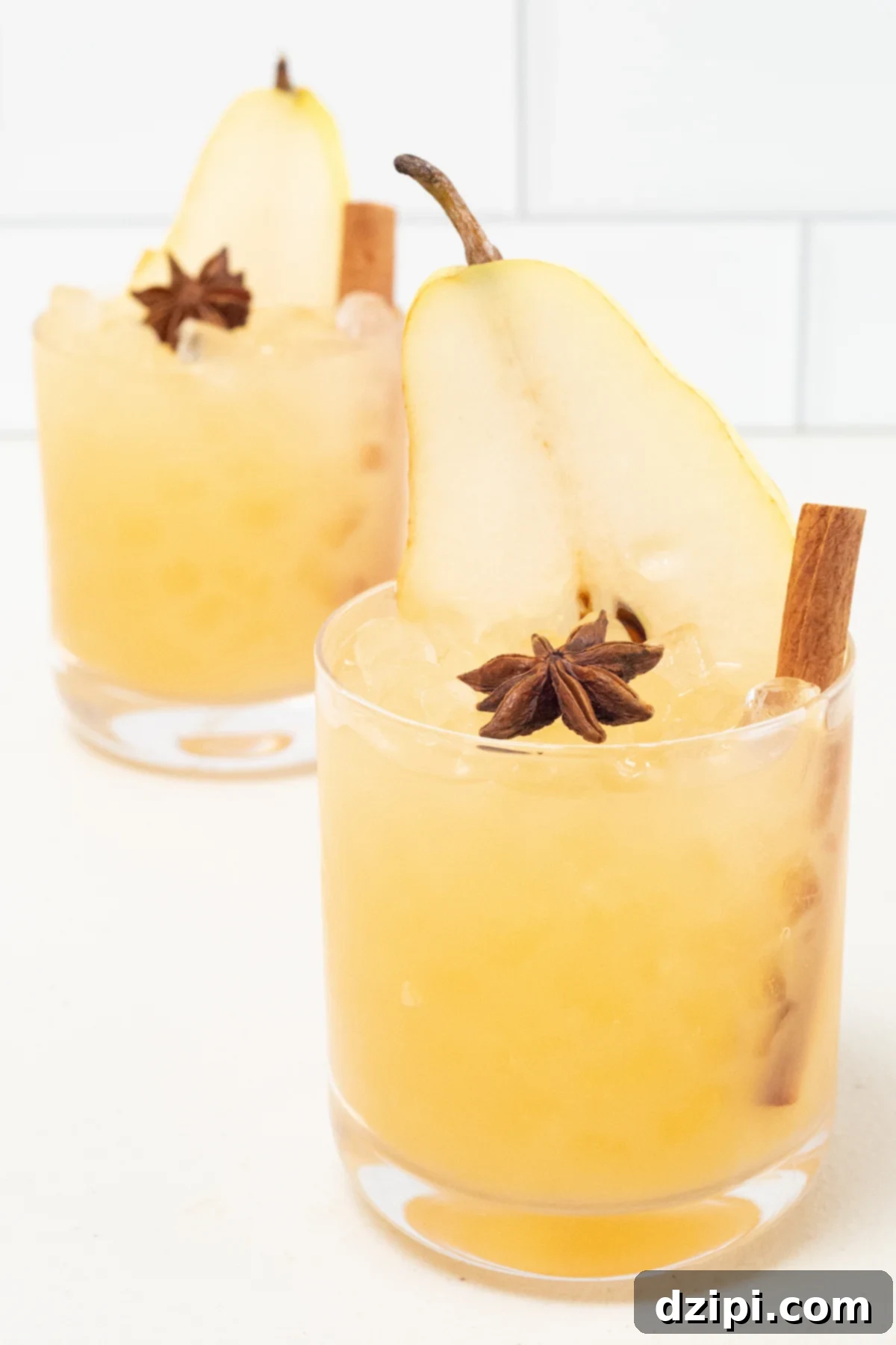 Two lowball glasses on a pristine white background, each holding a golden-yellow Thanksgiving cocktail. They are artfully garnished with a delicate pear slice, a fragrant star anise, and a slender cinnamon stick, showcasing elegant holiday presentation.