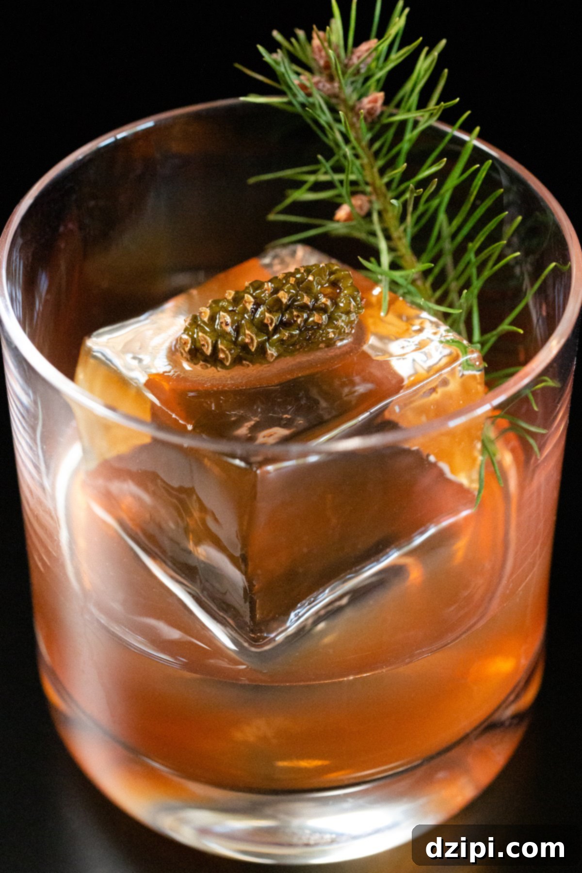 A festive Christmas Old Fashioned cocktail, garnished with a mini pine cone and a sprig of evergreen, sits in a lowball glass with a large clear ice block on a dark background. The reddish-brown liquid promises a taste of holiday cheer.
