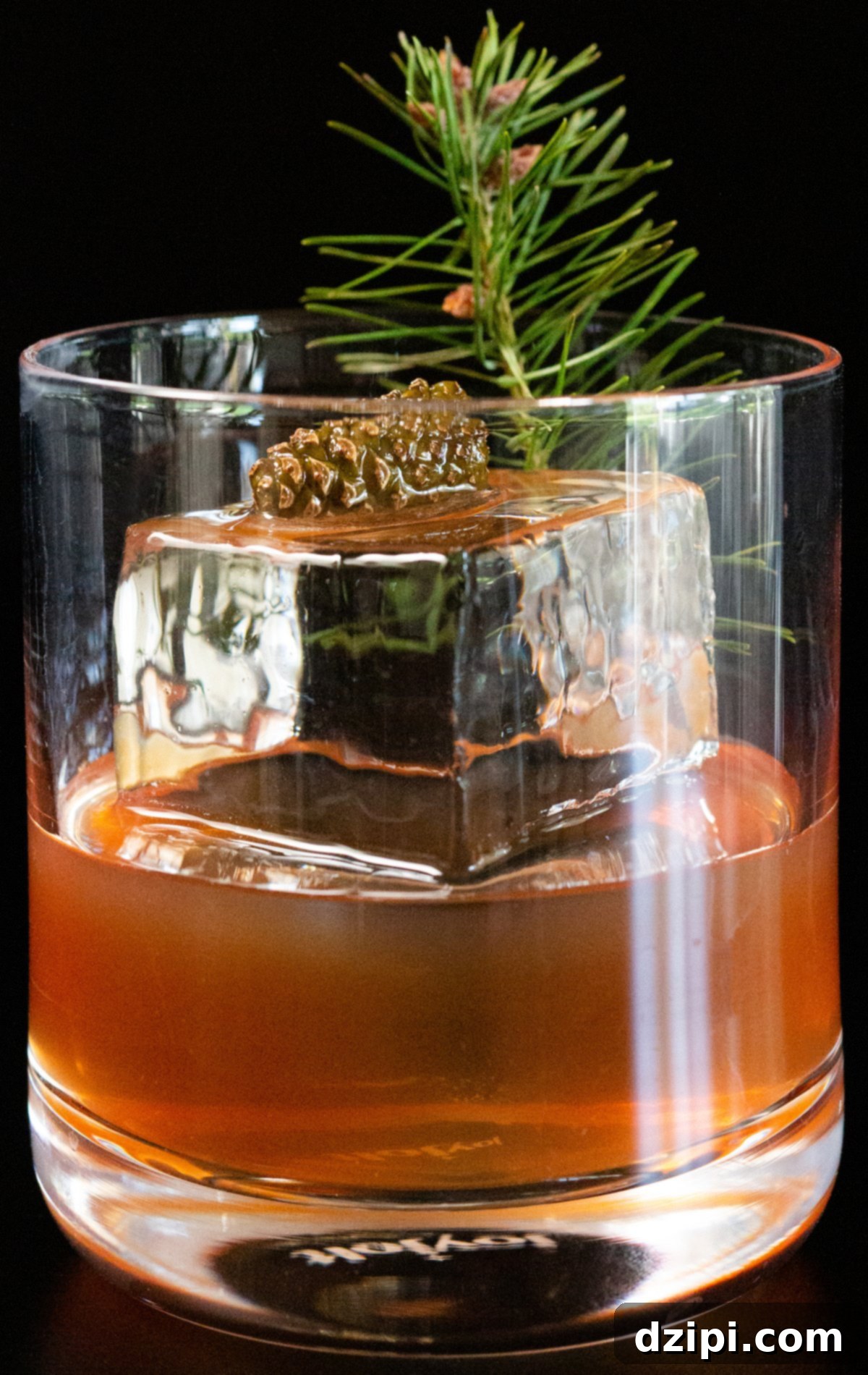 A beautifully presented Christmas Old Fashioned, showcasing a large ice block, fresh pine branch, and a tiny pine cone as garnishes, set against a dark, inviting background.