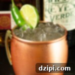 A copper mug filled with pebble ice and Mexican Mule in front of a tequila and chile liqueur bottle.