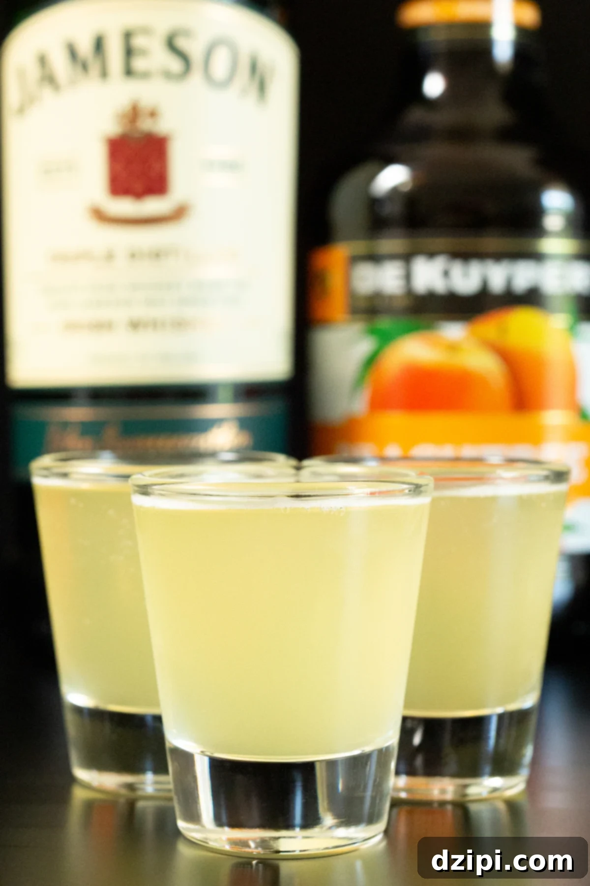 Three shot glasses filled with refreshing green tea shots, with bottles of Jameson Irish Whiskey and peach schnapps prominently displayed in the background.