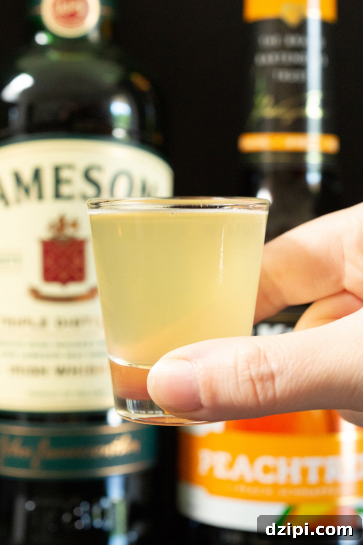 A hand gracefully holds up a clear shot glass, prominently featuring a vibrant and perfectly chilled Green Tea Shot, against a softly blurred background.