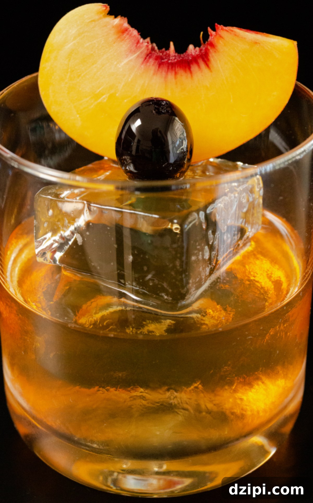 Close-up of a clear lowball glass with a large block of ice, partially filled with a Peach Old Fashioned, garnished with a peach slice and cherry.