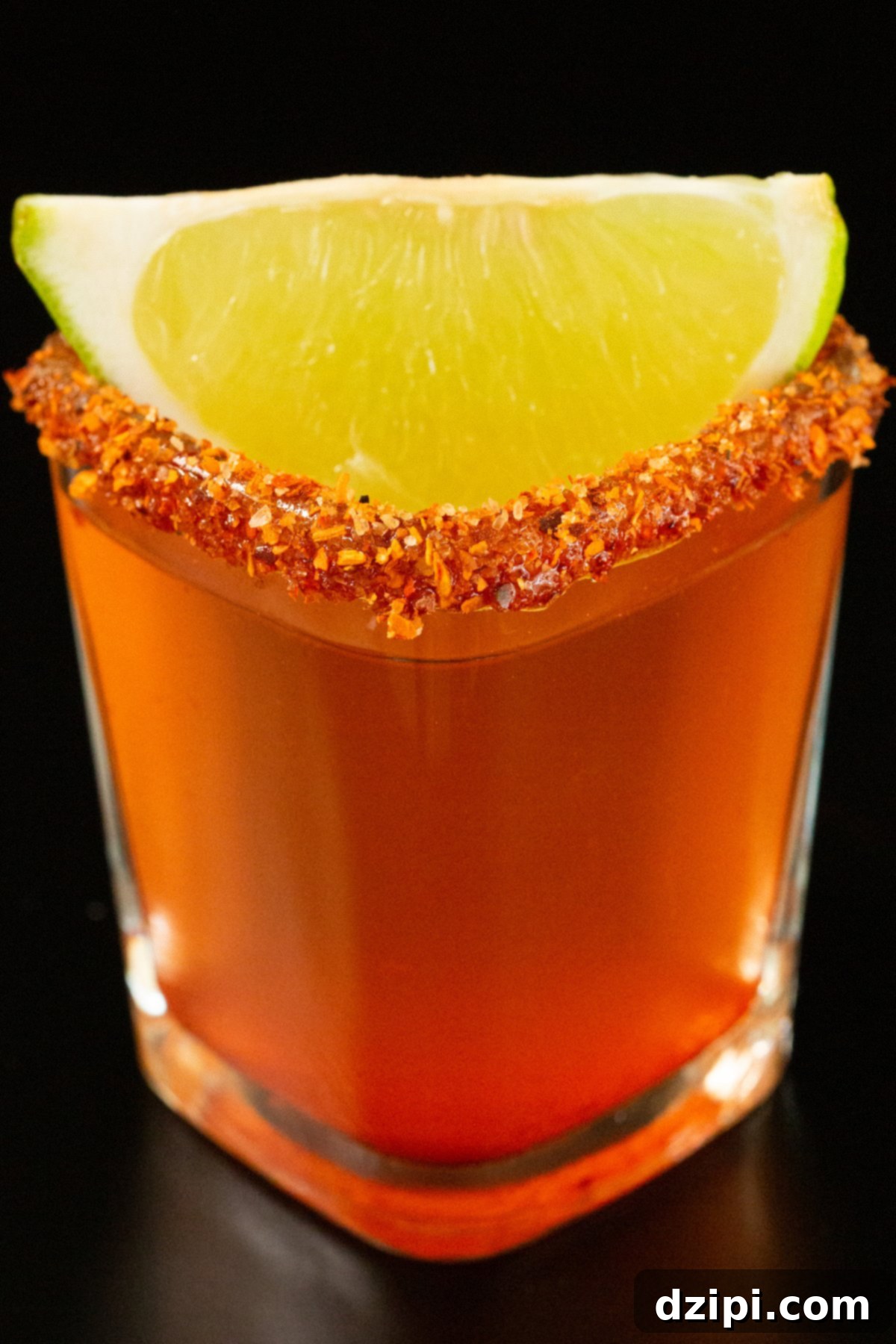 A single square shot glass has been filled with a pinkish-red Mexican candy shot. It is garnished with a chili powder rim and a lime wedge.