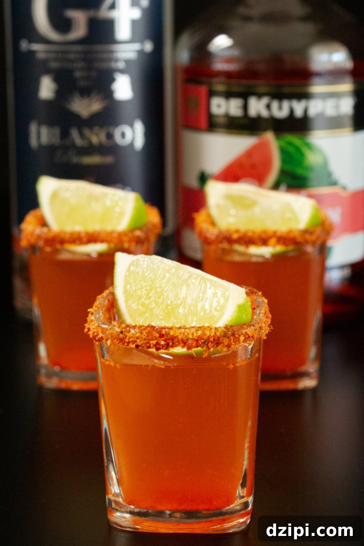 Three square Mexican Candy Shots in front of a bottle of tequila and watermelon schnapps.