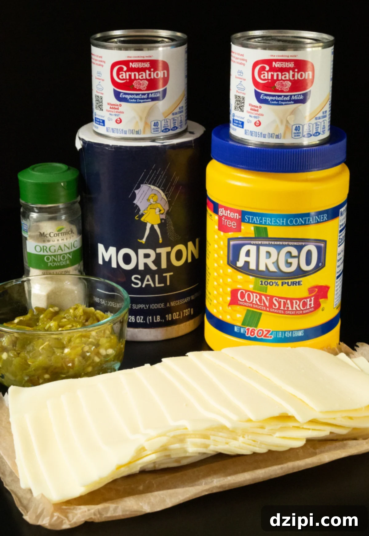A small prep dish of chopped hatch chiles and slices of white American cheese next to containers of cornstarch, salt, onion powder, and evaporated milk, laid out for a recipe.