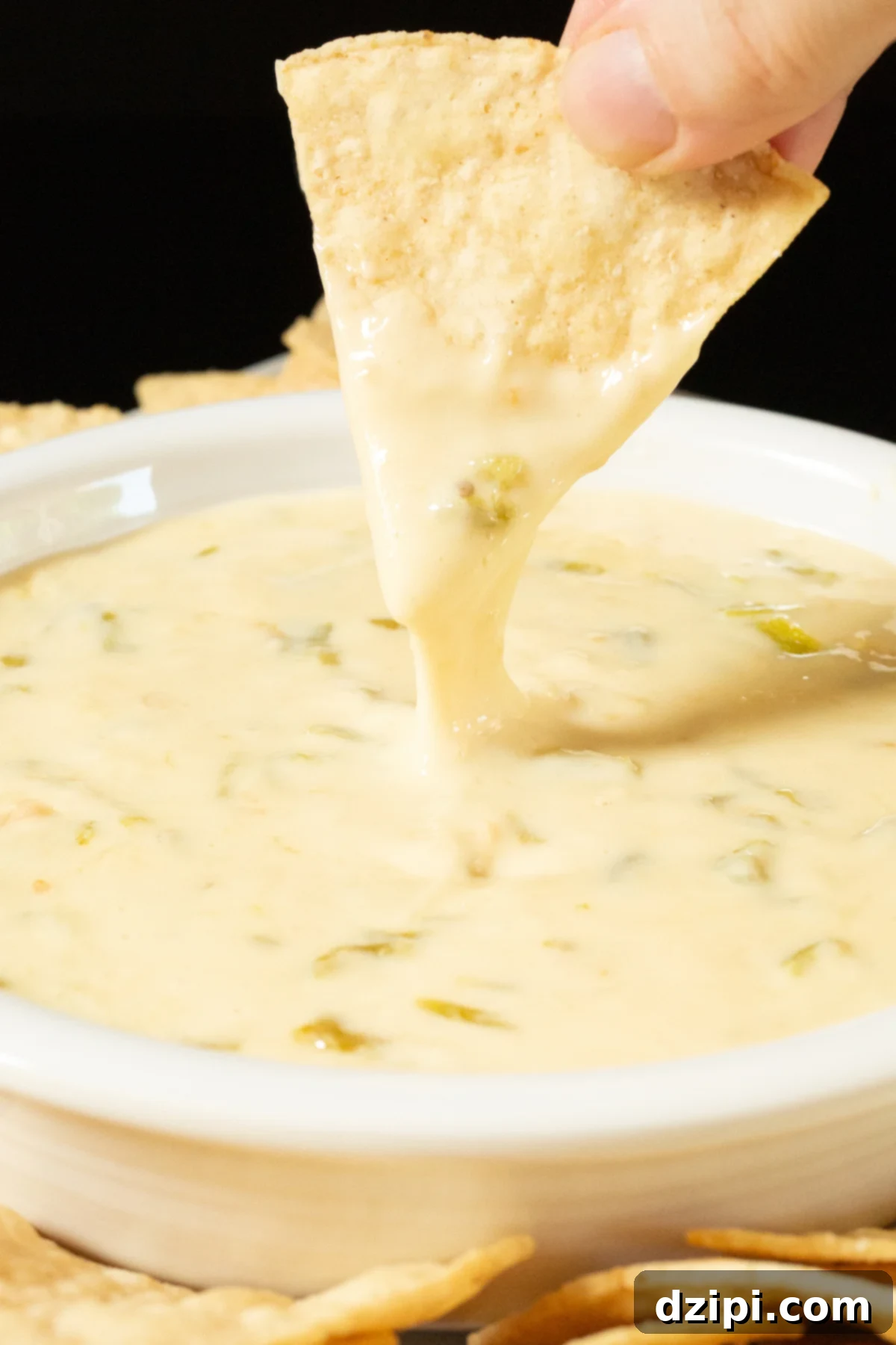 Two fingers pull a tortilla chip up from being dipped in a bowl of hatch chile queso, the cheese dip stretches with the chip in an appetizing manner.