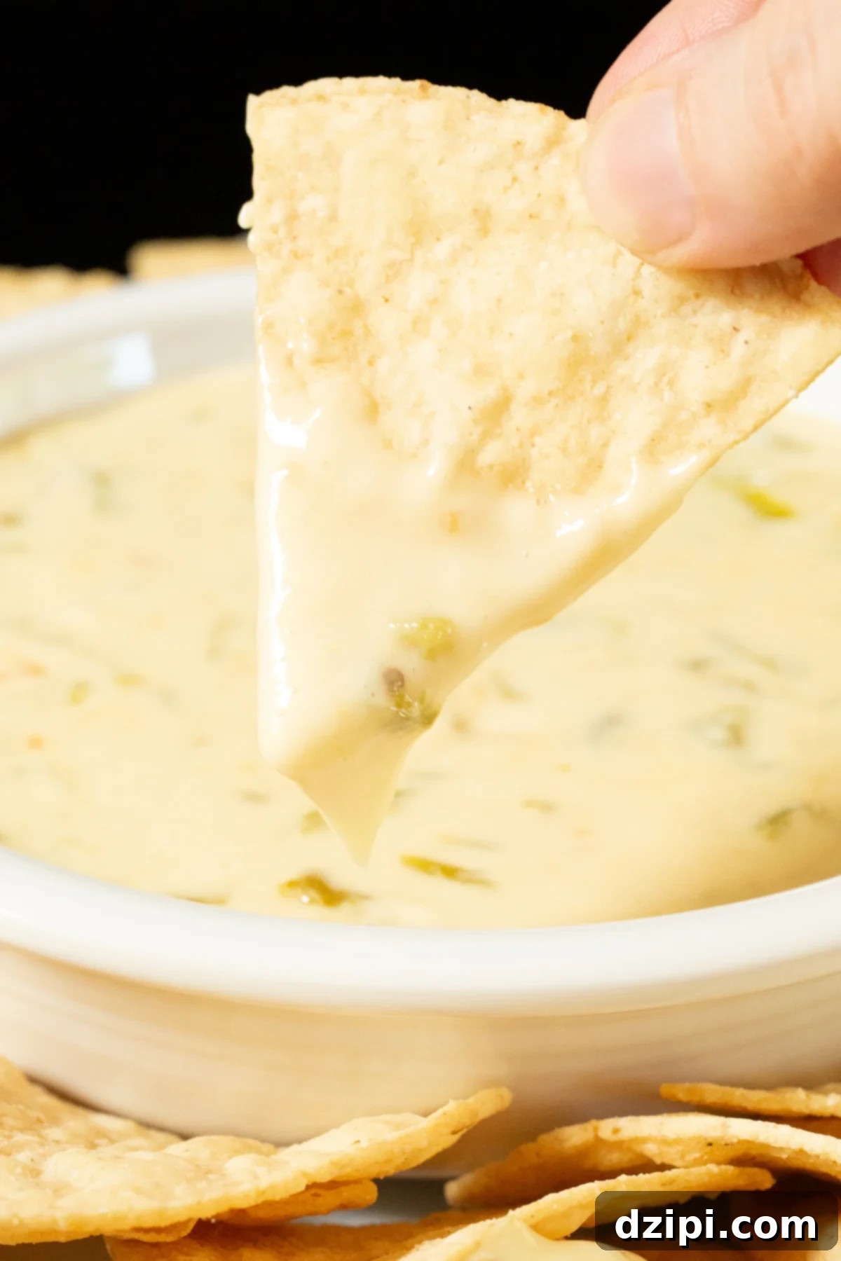 A hand holds up a tortilla chip that has been dipped in hatch chile queso, showing the creamy texture and stretch of the dip. A bowl with the rest of the dip is out of focus in the background.