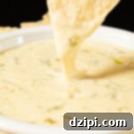 Two fingers pull a tortilla chip up from being dipped in a bowl of hatch chile queso, the cheese dip stretches with the chip.